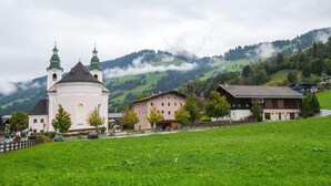 A church with two green domes, a pink building, and houses in a green valley with mountains in the background. - Apartment in Brixen Near Skiwelt Lift (Brixen im Thale)