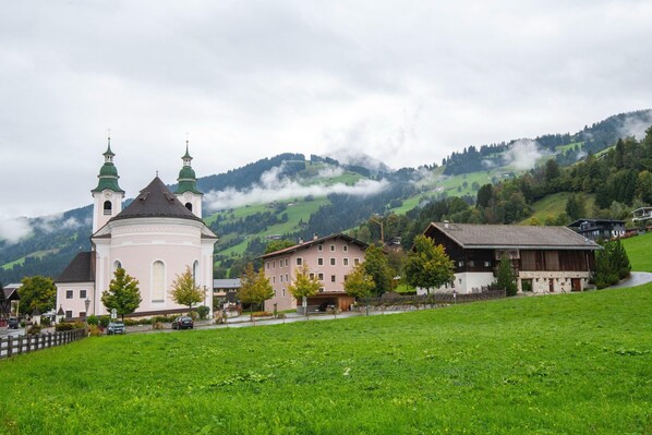 A church with two green domes, a pink building, and houses in a green valley with mountains in the background.