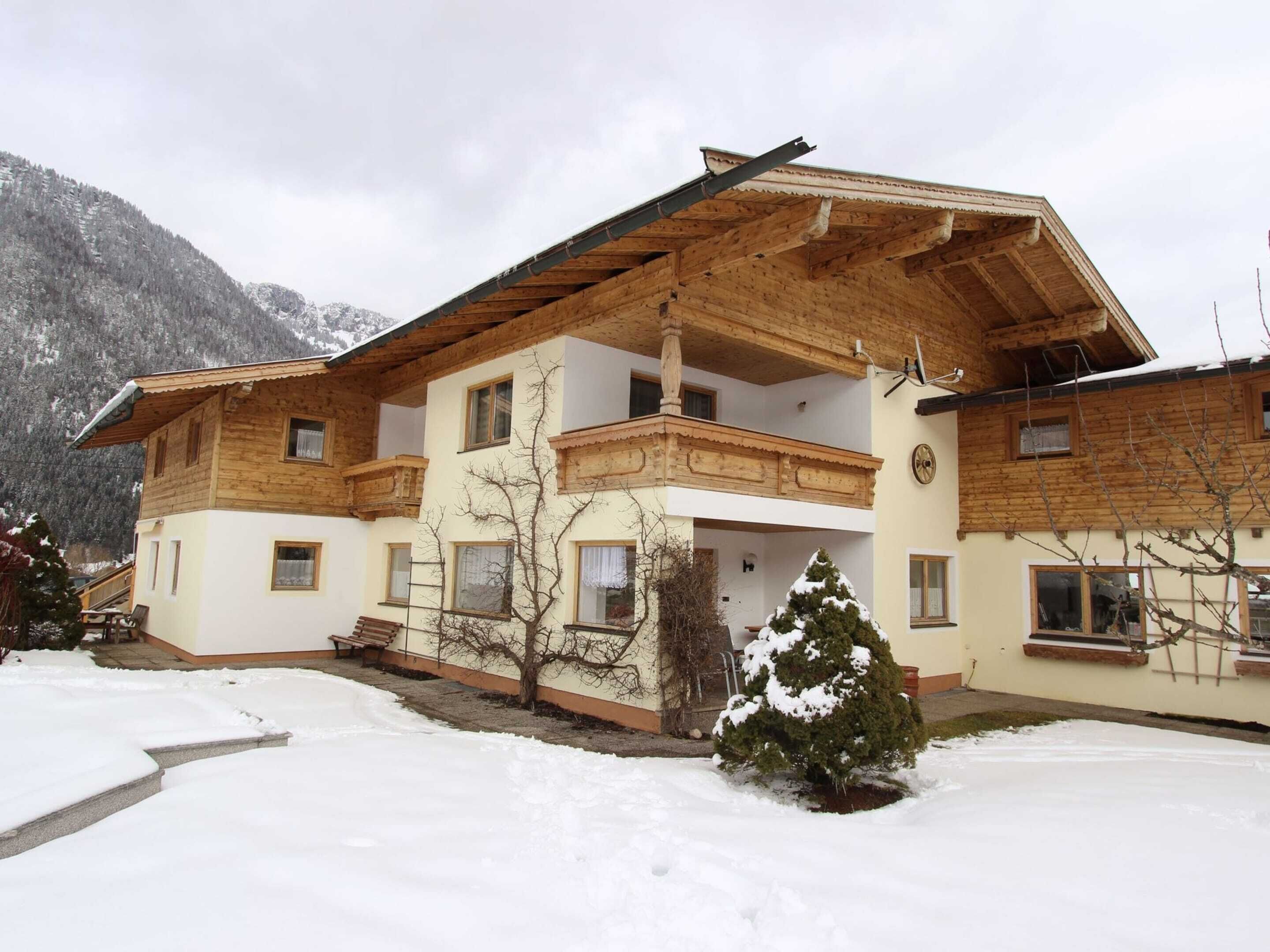 Un chalet en bois à deux étages, doté d’un balcon, entouré de terrain enneigé et avec des montagnes en arrière-plan.