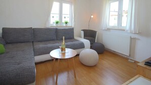 A modern living room with a grey sectional sofa, a round coffee table, and a small round ottoman. There are two windows with white curtains, a radiator, and a floor lamp. - Apartment in Brixen im Thale in ski Area (Brixen im Thale)