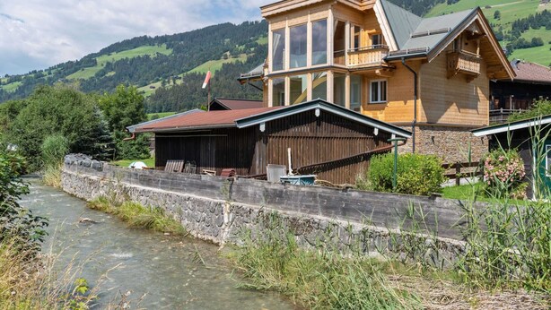 A wooden house with a stone wall and a river in the foreground. - Apartment in Brixen im Thale in ski Area (Brixen im Thale)