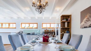 A dining area with a wooden table set for four, surrounded by grey chairs, a chandelier, and a bookshelf with books and decorative items. - Apartment With Sauna and Ski In/out (Mittersill)