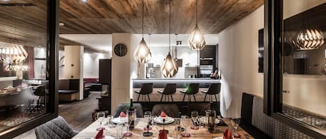 A modern dining area with a wooden table set for four, a kitchen in the background, and pendant lights above.