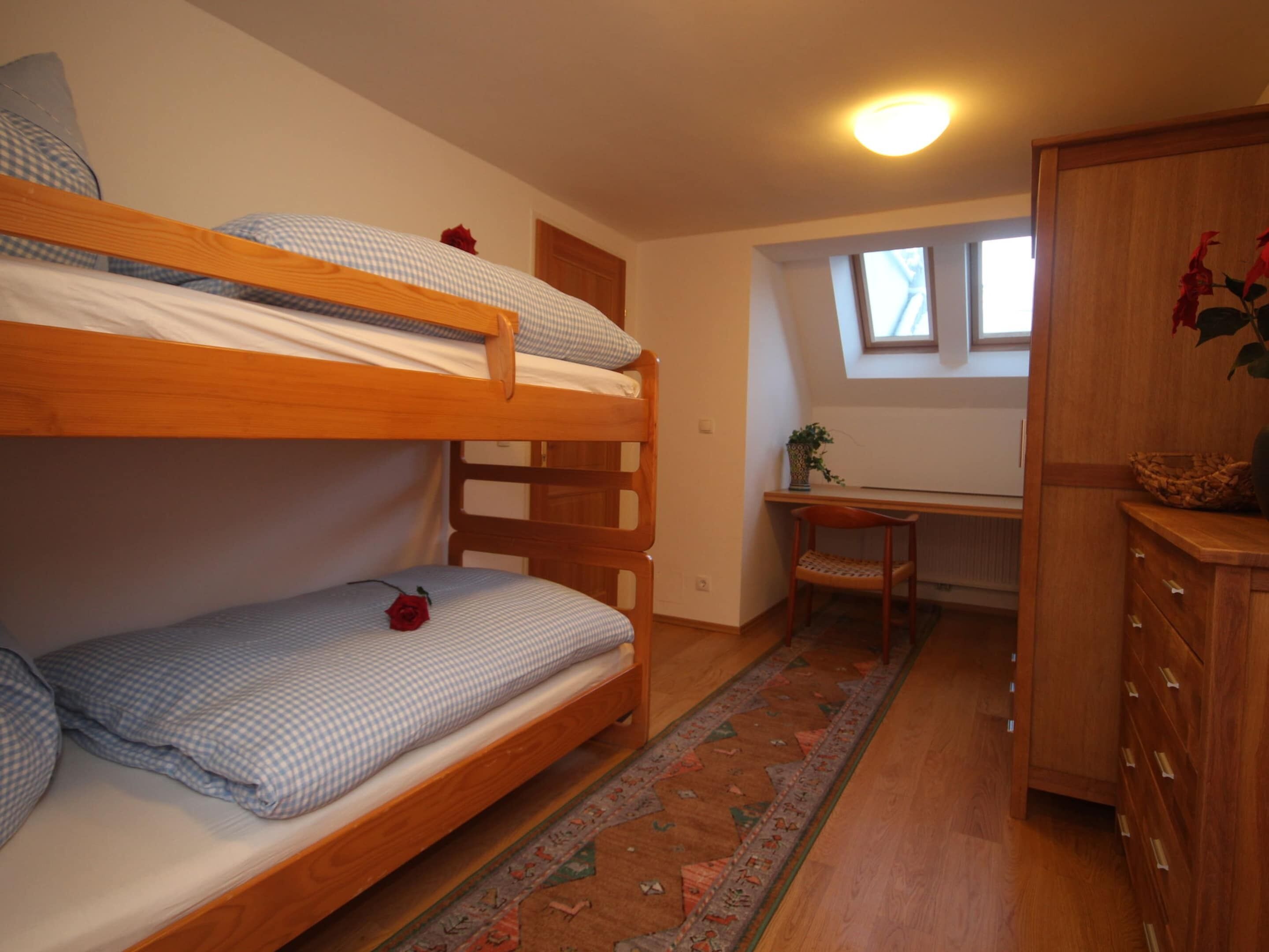 Ein Schlafraum mit Etagenbett, Schreibtisch, Stuhl, kleinem Fenster und Holzschrank.