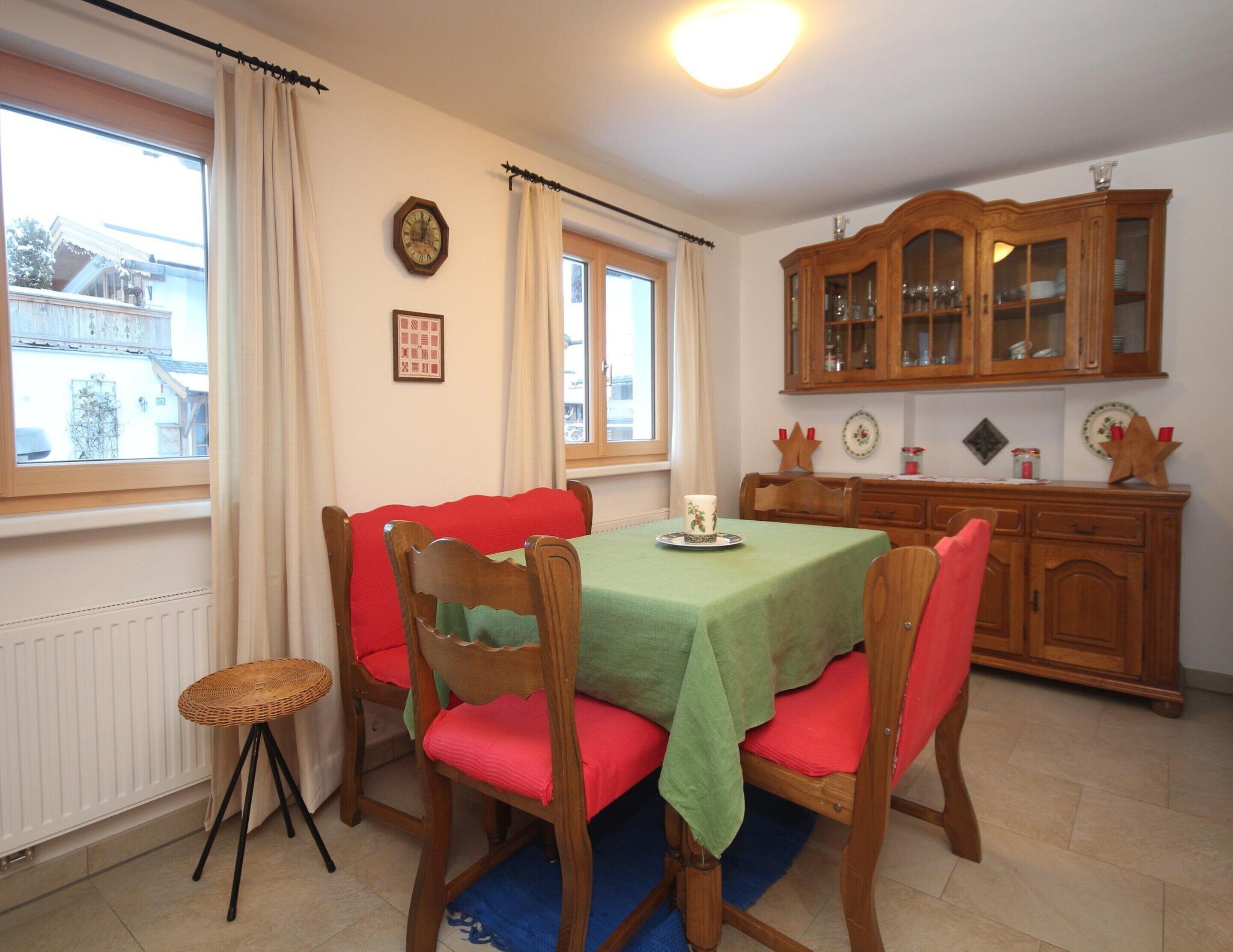 Un comedor con una mesa dispuesta para cuatro personas, rodeada de sillas de madera con cojines rojos. Un taburete de mimbre está colocado al lado de la mesa.