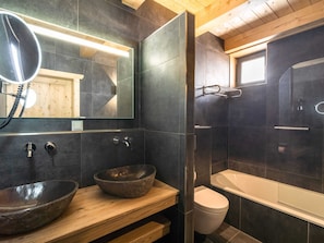A modern bathroom with dark tiles, a wooden vanity, a large mirror, a round vessel sink, a toilet, and a walk-in shower.