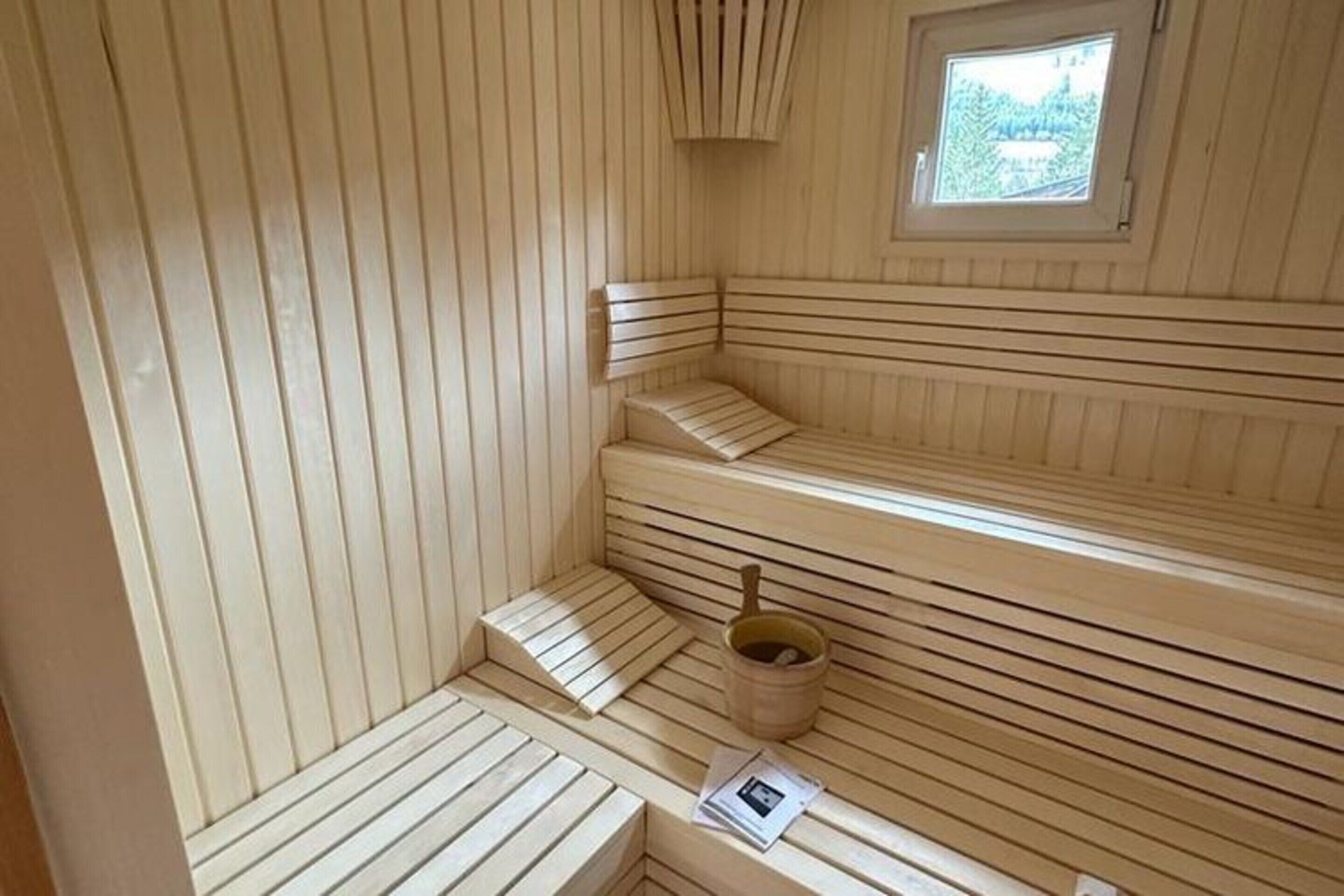 Una sauna con varias bancas, un cubo de madera y un ventanal pequeño.