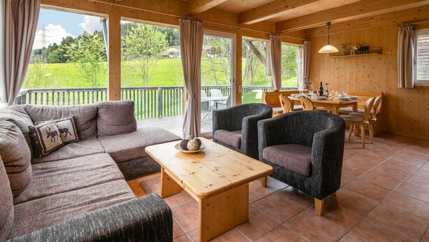 A cozy cabin interior with a wooden ceiling, a sofa, two armchairs, a dining table with chairs, and large windows offering a view of a green landscape. - Wooden Chalet in Styria near Kreischberg Ski Area (Sankt Georgen am Kreischberg)