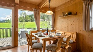 A wooden dining room with a table set for four, chairs, a hanging light fixture, and a view of a green landscape through large windows. - Wooden Chalet in Styria near Kreischberg Ski Area (Sankt Georgen am Kreischberg)