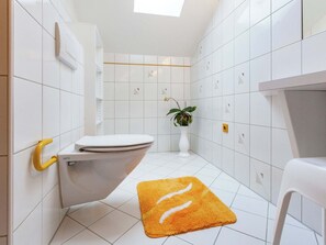 A modern bathroom with white tiles, a toilet, a sink, a plant, and a yellow rug.