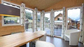A wooden interior with a dining table and chairs, large windows with curtains, and a view of a wooden house outside. - Wooden Chalet in Hohentauern With Sauna (Hohentauern)
