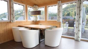 Una cabaña de madera con zona de comedor, que incluye una mesa y cuatro sillas blancas. Amplias ventanas ofrecen vistas a una zona montañosa boscosa.