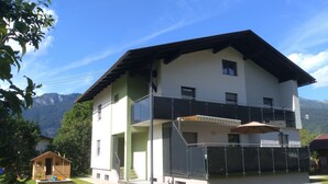 A two-story building with a balcony, a small wooden structure nearby, and mountains in the background.
