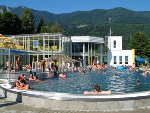 A swimming pool area with people enjoying the water, a building with large windows, and a mountainous backdrop.