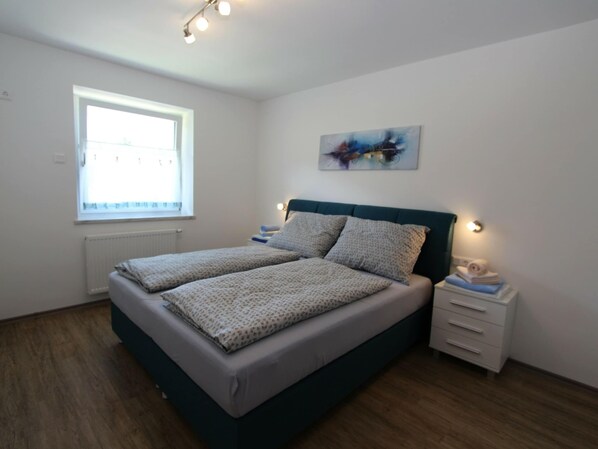 A bedroom with a bed, a window, a radiator, a nightstand, and a wall-mounted light fixture.