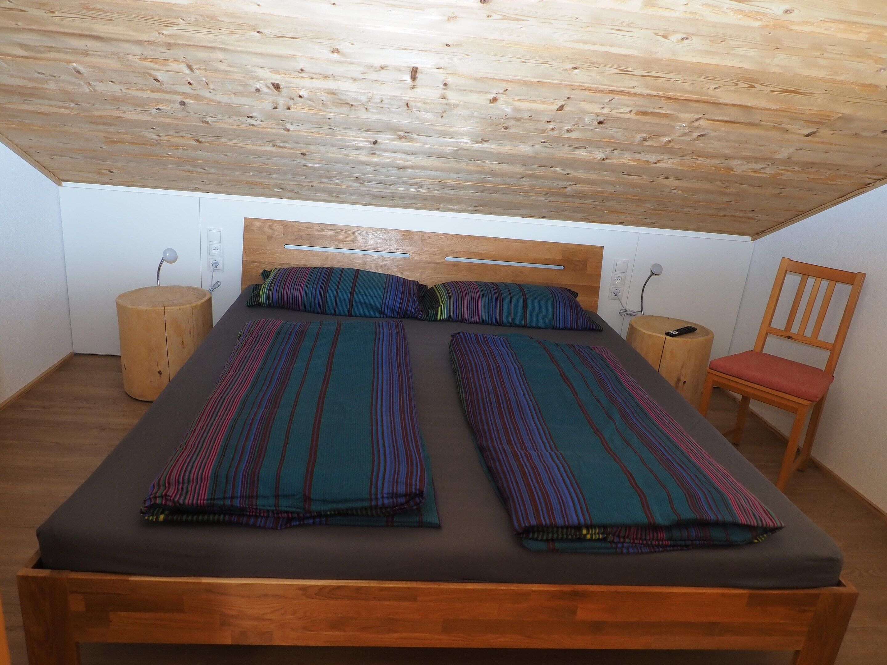 Una stanza accogliente e piccola con soffitto in legno, un letto con lenzuola a righe, una sedia in legno e un piccolo sgabello di legno.