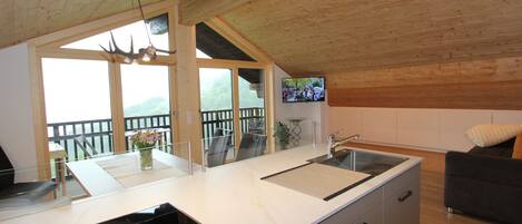 A modern kitchen with a large island, a built-in stove, and a sink. There is a balcony with a view of the mountains.