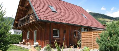 A wooden cabin with a red-tiled roof, a wooden deck, and a pond in the foreground.