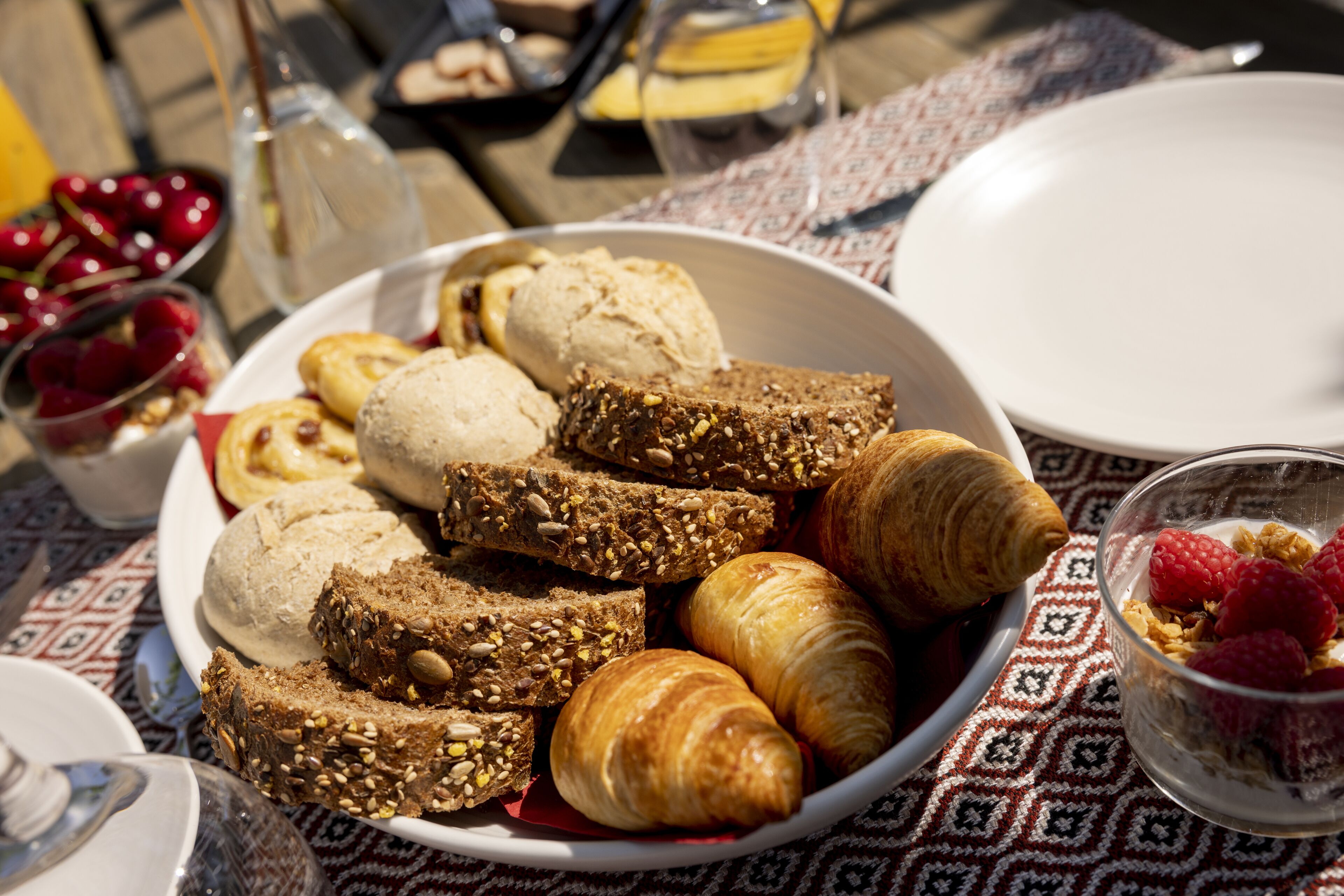 Une table mise pour le petit-déjeuner, avec des croissants, du pain et un saladier de fruits sur une nappe à motifs.
