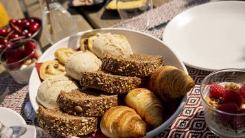 Une table mise pour le petit-déjeuner, avec des croissants, du pain et un saladier de fruits sur une nappe à motifs.