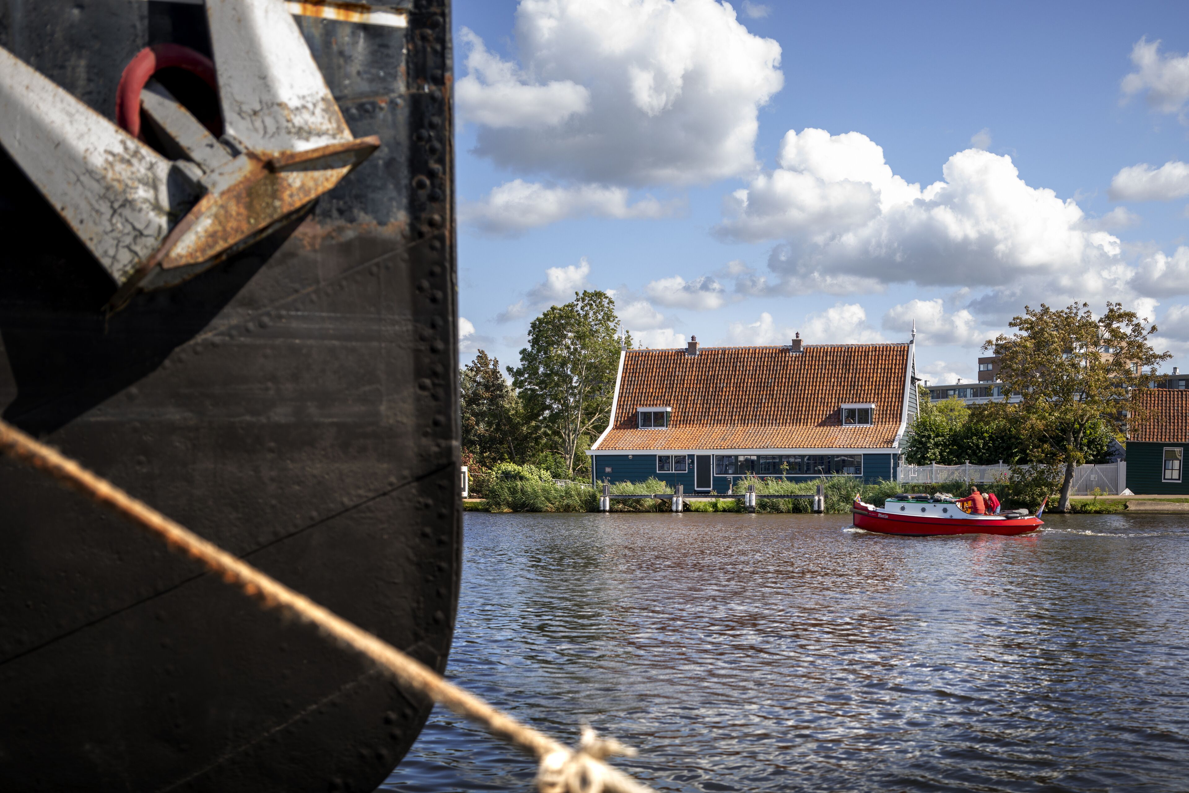 A boat with a red and white cabin, docked near a house with a red-tiled roof, surrounded by trees and a clear sky.