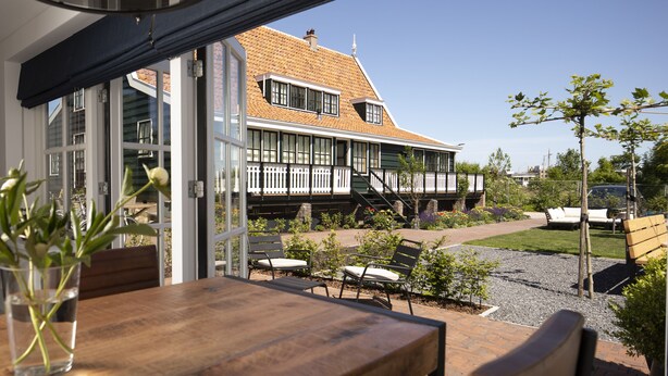 An outdoor seating area with a wooden table and chairs, a potted plant, and a view of a house with a sloped roof and white railings. - B&B Saenliefde (Wormer)