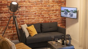 A living room with a brick wall, a dark grey sofa, a coffee table with wine glasses and a bottle, and a mounted TV displaying zebras. - B&B Saenliefde (Wormer)