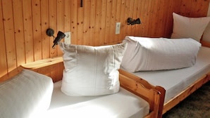 Une chambre dans un chalet en bois, avec deux lits simples, un banc en bois et une croix au mur.