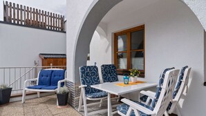 Ein Sitzbereich im Freien mit weißen Möbeln und blauen Kissen mit Muster, ein kleiner Tisch mit einer Laterne und ein Fenster mit Holzläden.