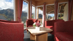 Una habitación con sillones tapizados en rojo, una mesa de centro de madera con un florero y grandes ventanales con vistas a la montaña.
