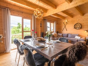 A wooden interior with a dining table set for four, a sofa, and a view of greenery outside. - Cozy Holiday Home in Finkenstein near Lake (Finkenstein am Faaker See)