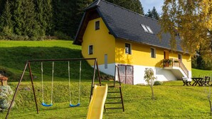 Una casa gialla con tetto nero, altalene e uno scivolo in un'area erbosa.