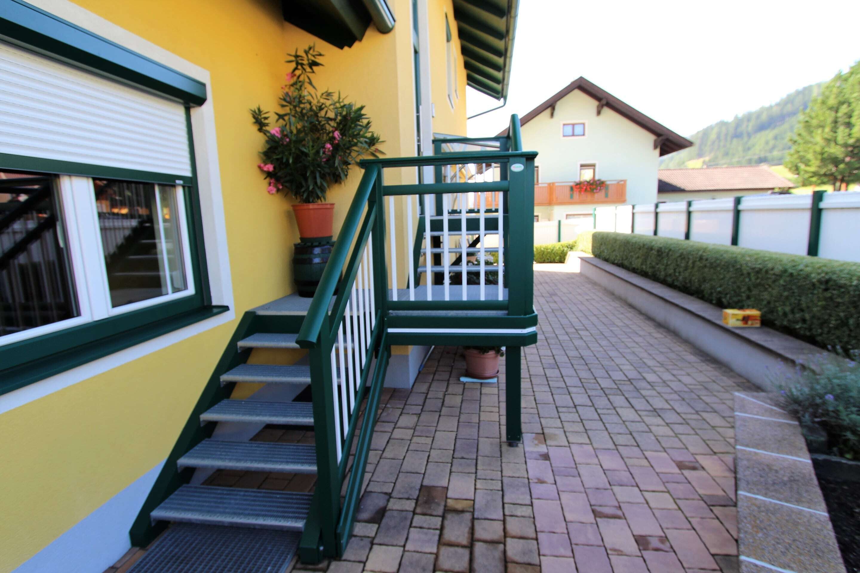 Un bâtiment jaune avec un escalier vert menant à une porte, un pot de fleurs et une haie.