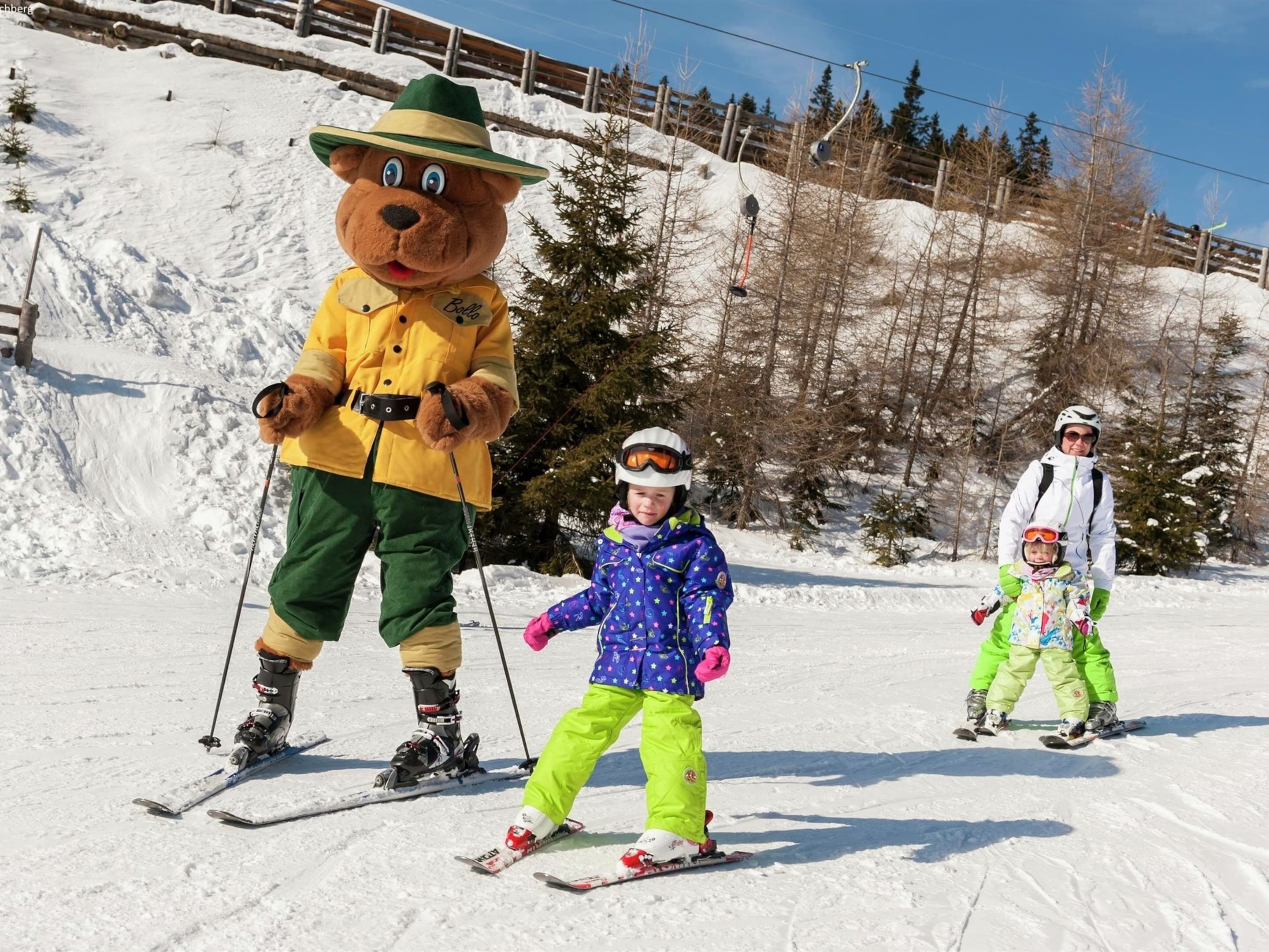 A mascot on skis, a child in a blue jacket, and an adult in a white jacket on a snowy slope.
