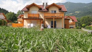 Une maison à deux étages avec un balcon, entourée d’un champ de maïs et avec des montagnes en arrière-plan.