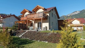 Une maison à deux étages dotée d’un balcon, entourée d’un jardin bien entretenu et avec des montagnes en toile de fond.