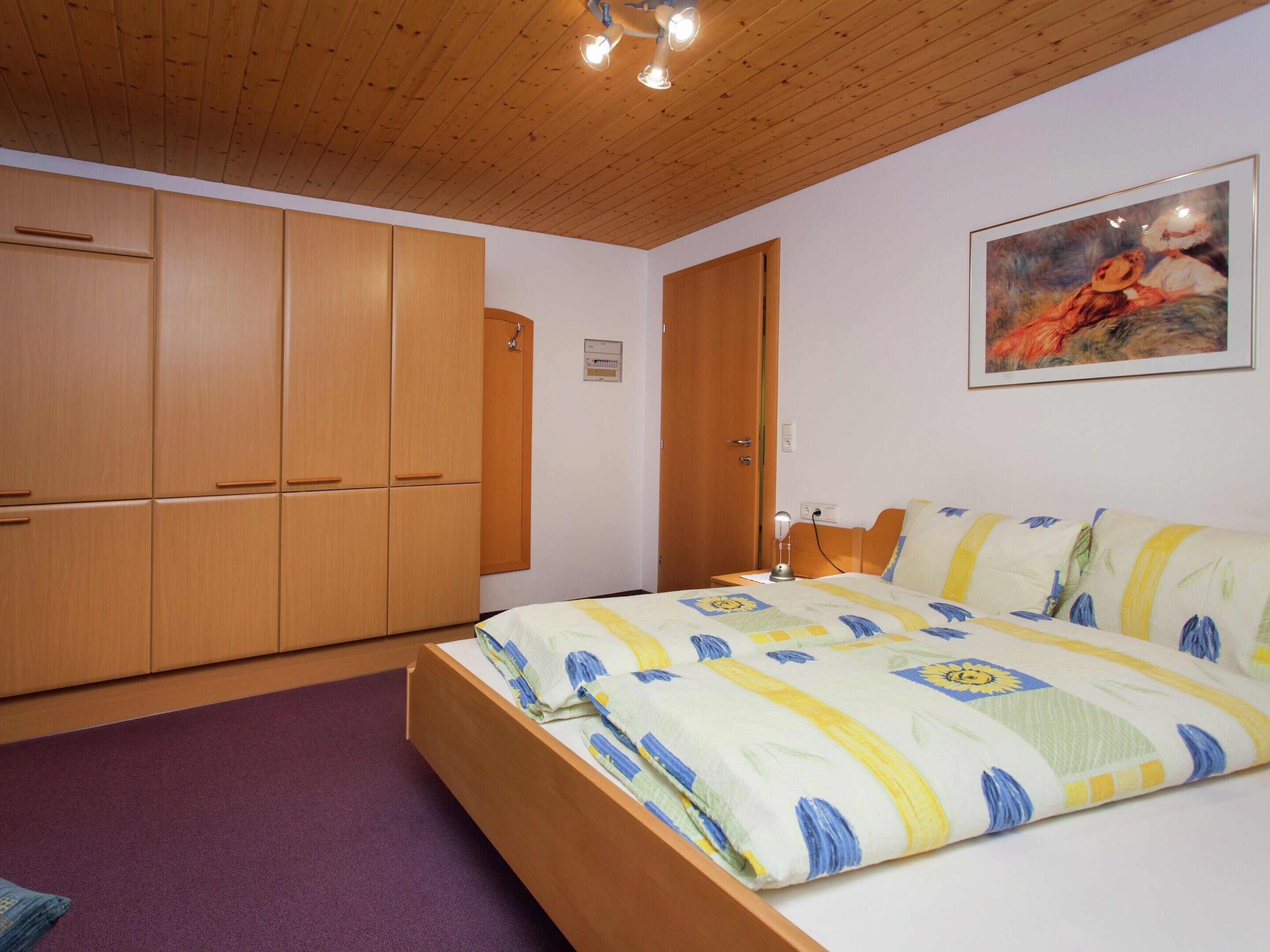 A bedroom with a wooden ceiling, a bed with a patterned bedspread, a nightstand with a lamp, and a painting on the wall.