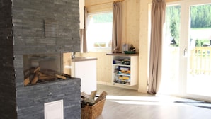 A modern living room with a stone fireplace, a wooden bookshelf, and a large window with curtains. - Wooden Chalet in Hohentauern With Sauna (Hohentauern)