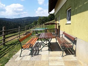 A patio with wooden benches and a table, a yellow building with a window, and a view of a forested mountain range. - Chalet in Sankt Andra Near Koralpe Ski Area (Sankt Andra)