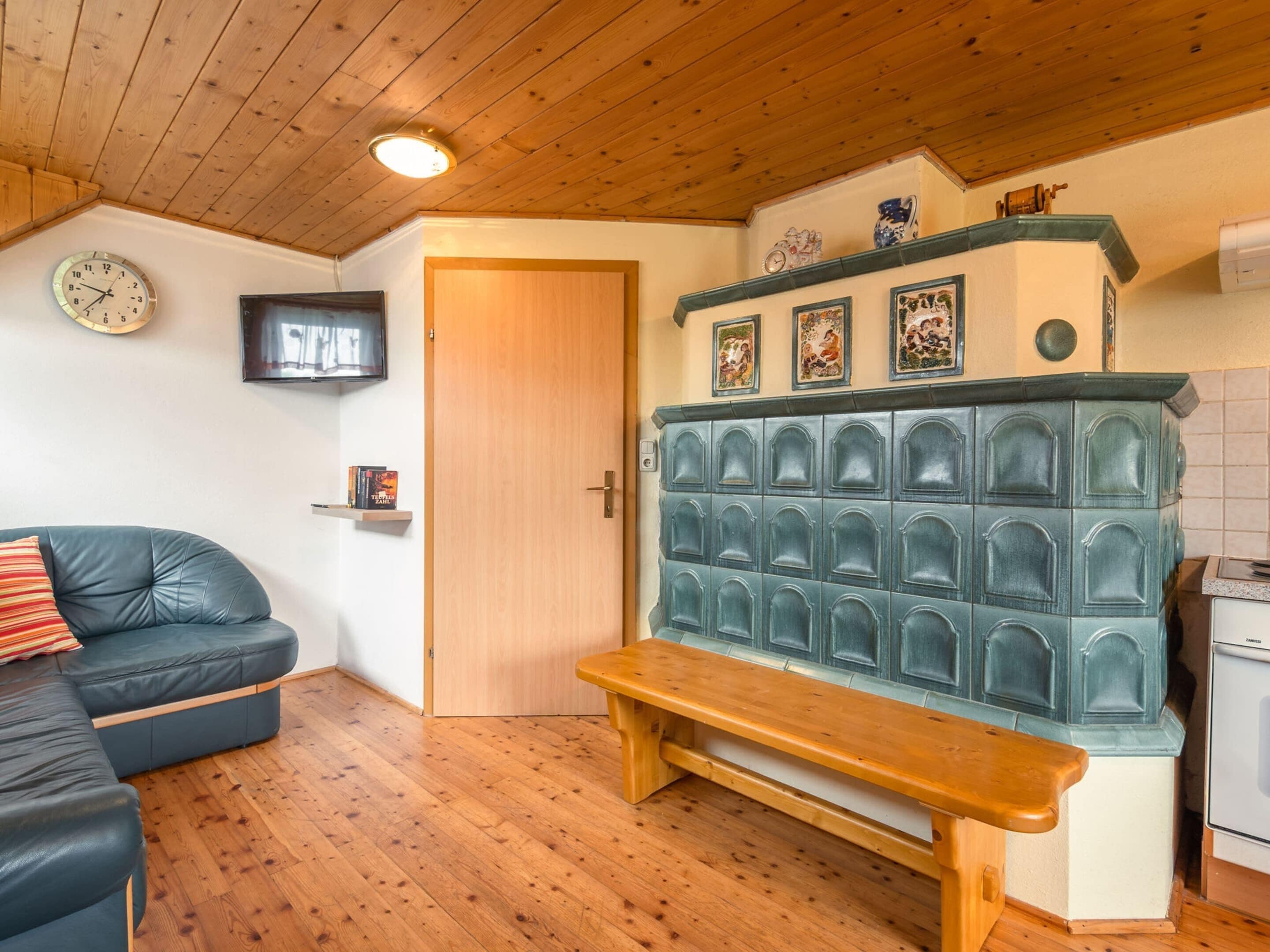Um quarto aconchegante com teto de madeira, um sofá azul, um banco de madeira, uma TV fixada na parede e um armário decorativo com obras de arte.