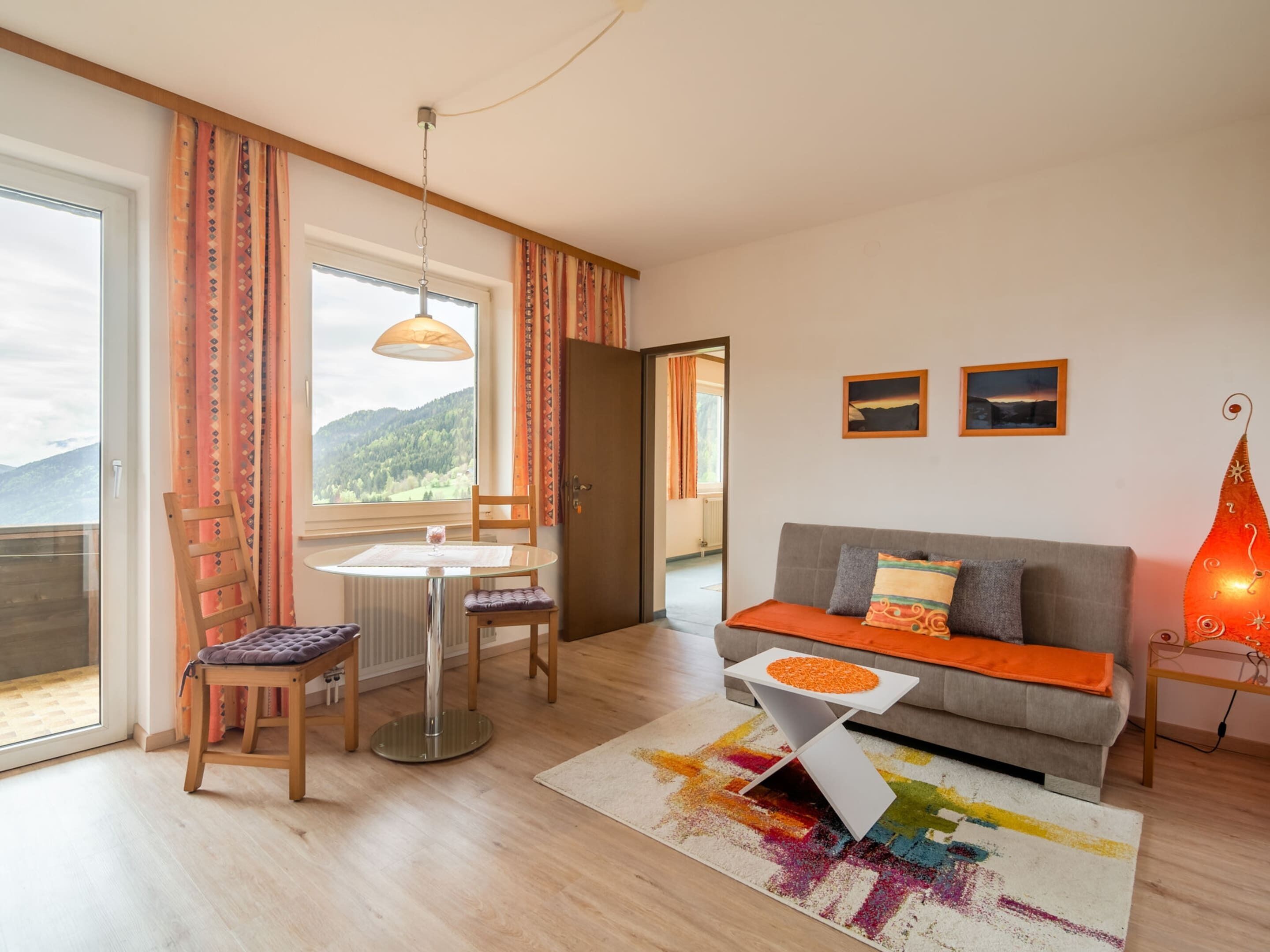 A cozy living room with a sofa, a small table, and a chair. There is a window with curtains, a radiator, and a view of the outdoors.