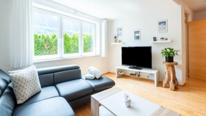 A modern living room with a sectional sofa, a flat-screen TV, wooden flooring, and a large window offering a view of greenery. - Apartment in Strengen Near Ski Bus (Strengen)