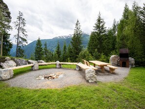 A rustic outdoor seating area with stone benches and a fire pit, surrounded by a natural landscape with mountains in the background.