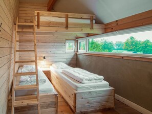 A wooden cabin room with bunk beds, a ladder, a window, and a small desk with a lamp.