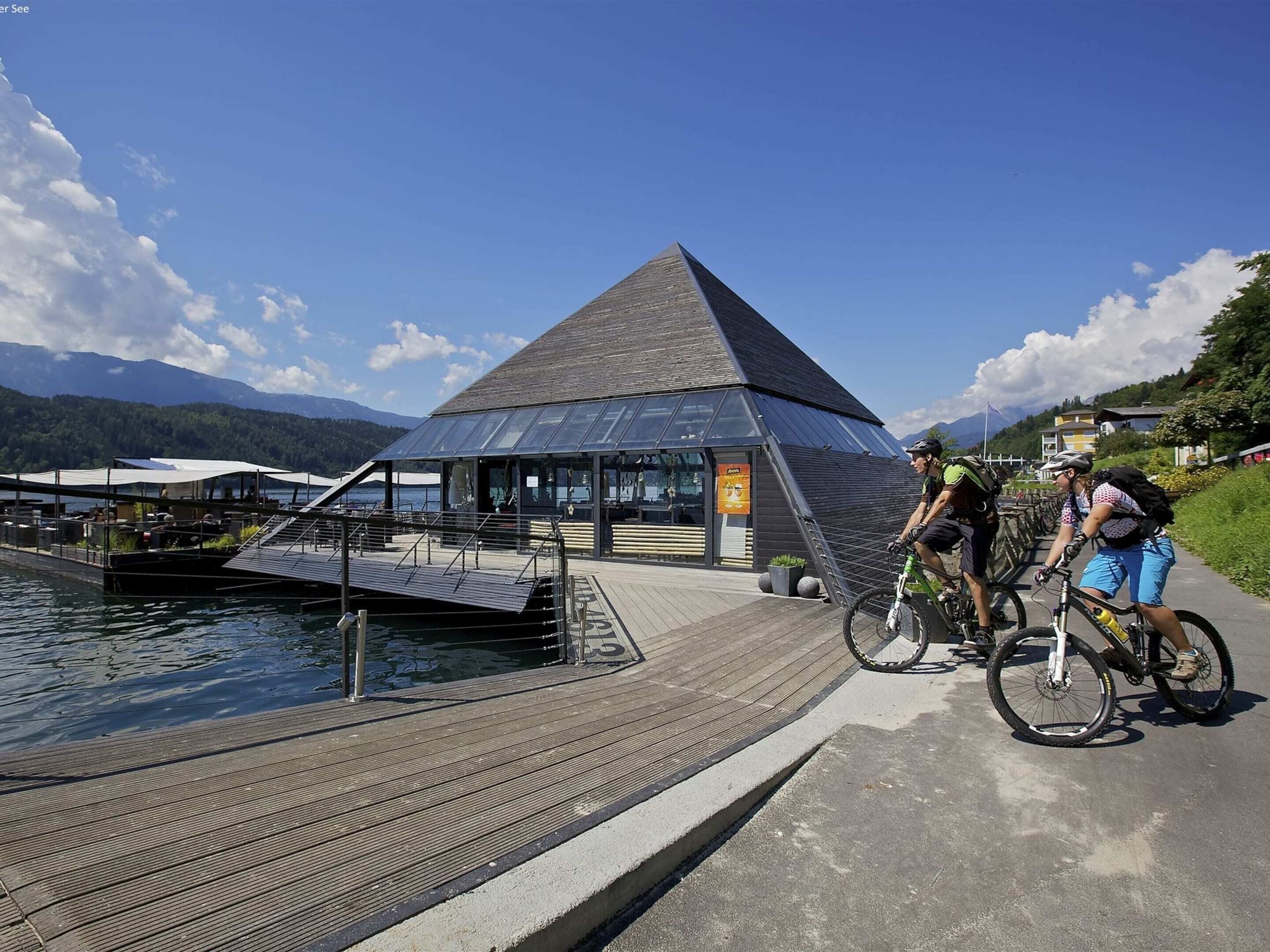 A pyramid-shaped building with a glass facade, a dock extending into a lake, and two cyclists on a nearby path.