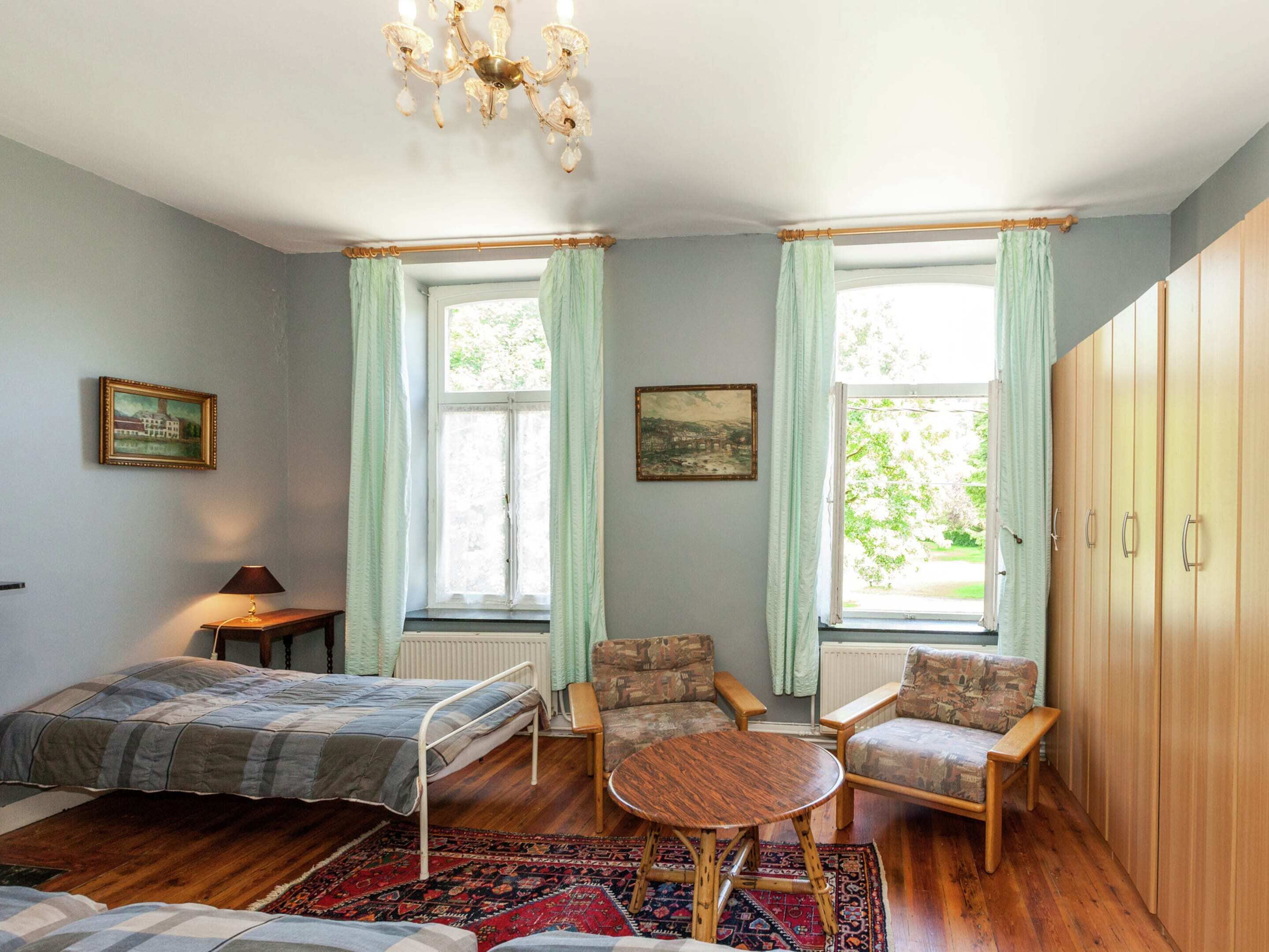 Ein Zimmer mit zwei Betten, einem Holzschrank, einem Tisch, zwei Stühlen und zwei Fenstern mit Vorhängen.