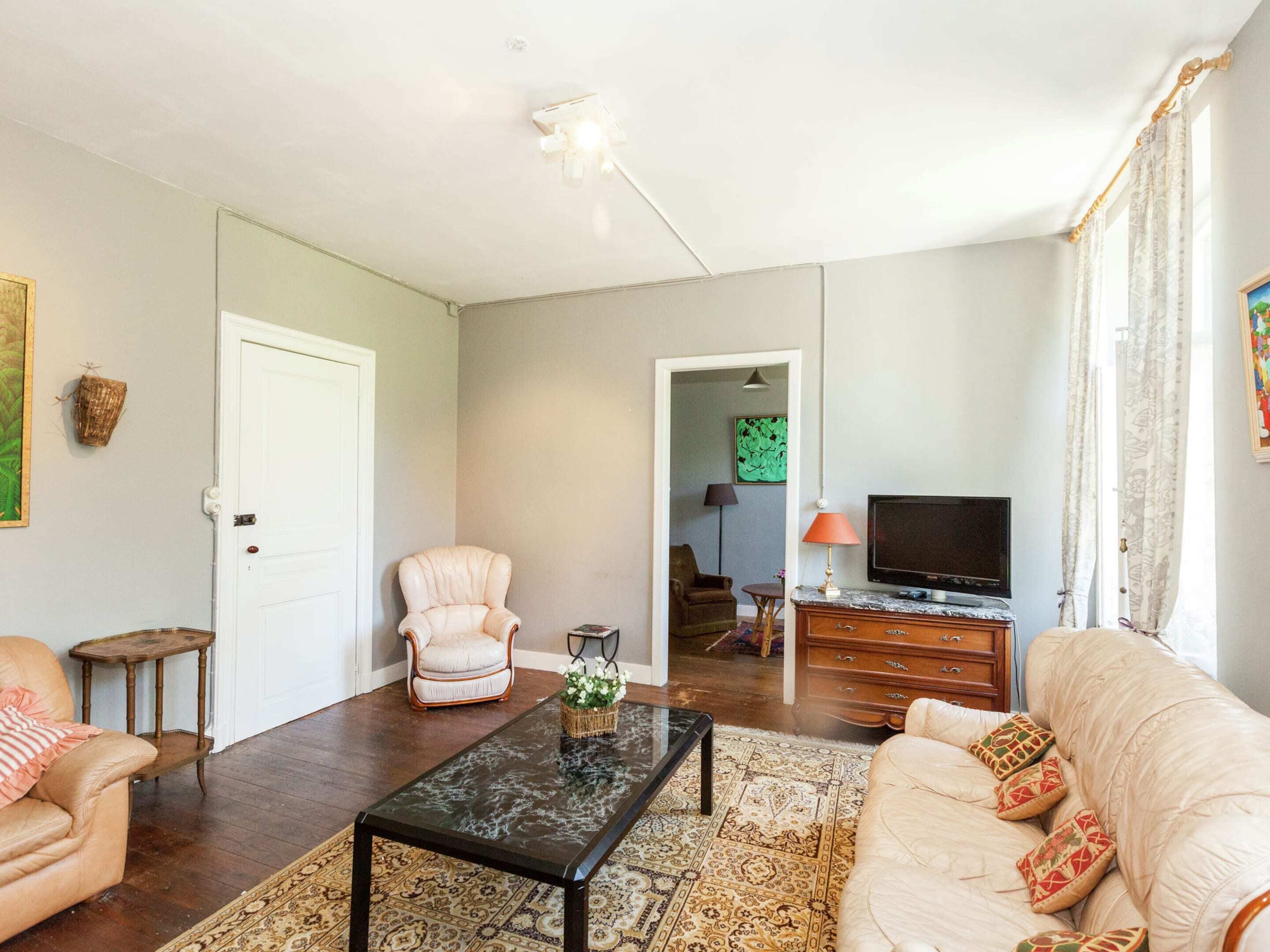 A living room with a sofa, armchair, coffee table, and TV. There is a door, a small table, and a patterned rug.