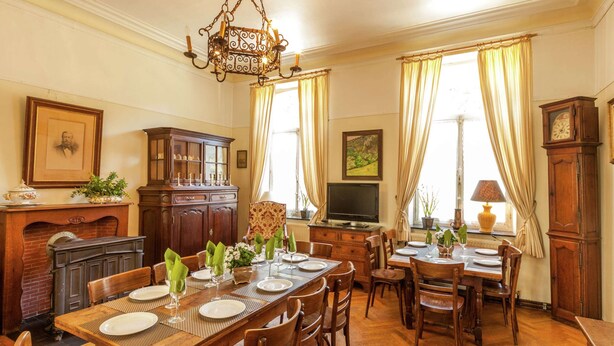 A dining room with a large wooden table set for six, flanked by two smaller tables. A television is mounted on the wall, and a grandfather clock is visible to the right.