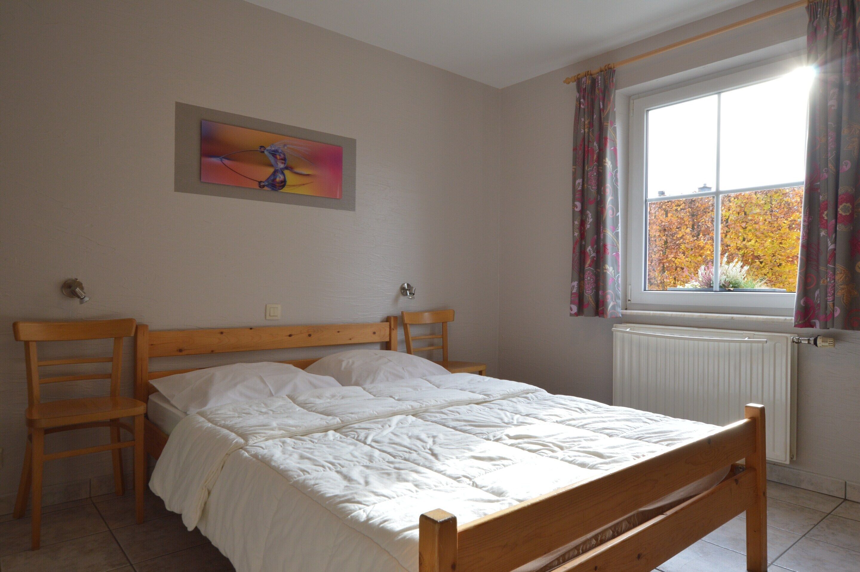 A bedroom with a wooden bed, two wooden chairs, a radiator, a window with curtains, and a painting on the wall.