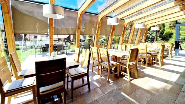 A sunlit dining area with wooden tables and chairs, large windows, and a view of the outdoors. - Apartment in Bad Kleinkirchheim (Bad Kleinkirchheim)